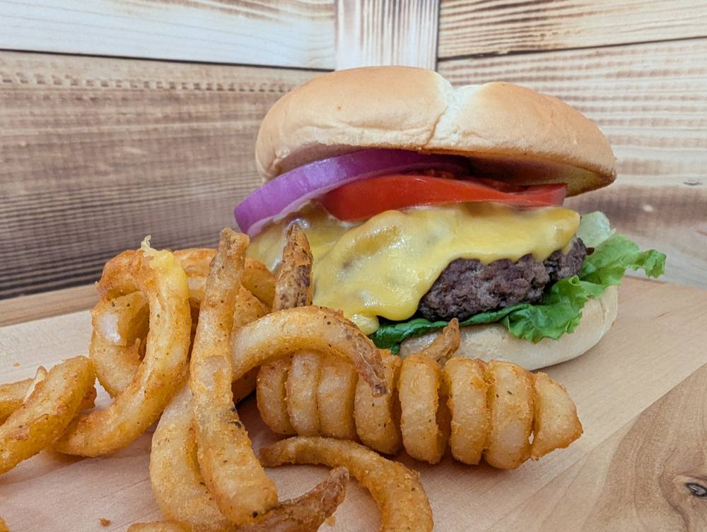 A cheeseburger on a toasted bun with lettuce, tomato and red onion, served with a pile of seasoned curly fries.