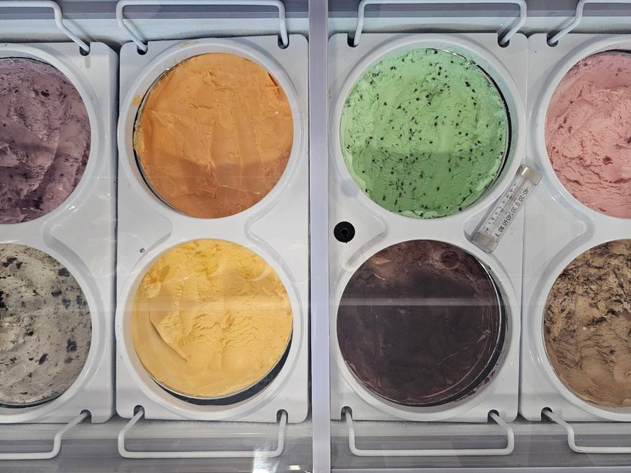 Eight open tubs of ice cream in the dipping cabinet, including huckleberry, mint chip, strawberry and cookies-and-cream.