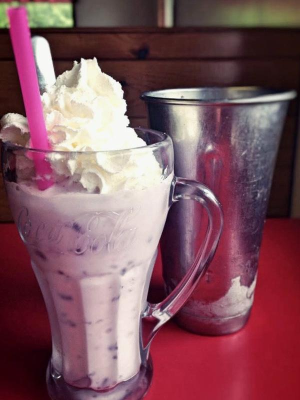 A huckleberry milkshake in a Coca-Cola fountain glass topped with whipped cream, next to the metal mixing tin it was spun in.
