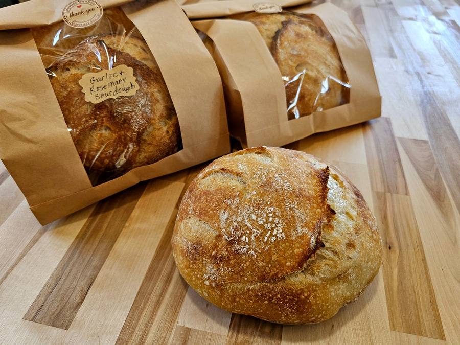 Two bagged sourdough loaves with handwritten labels reading Garlic and Rosemary Sourdough, beside a bare golden loaf on a butcher block.
